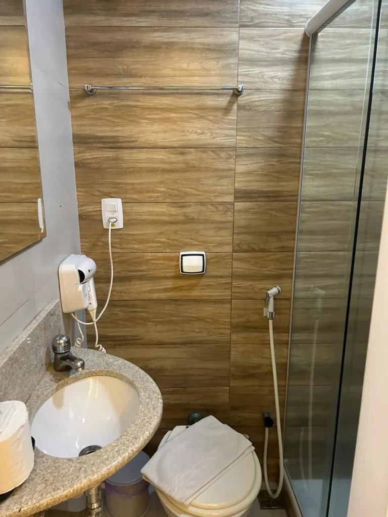 Banheiro da minissuíte com revestimento amadeirado, box de vidro, secador de cabelo e ducha higiênica