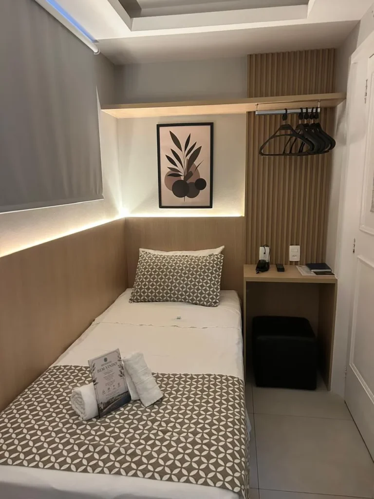 Suíte Cápsula (Mini Suíte) reformada, com cama de solteiro, painel de madeira ripado, cabideiro e mesinha com pufe