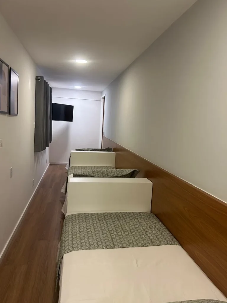 Suíte Cápsula tripla com três camas em fila, painel de madeira e TV na parede