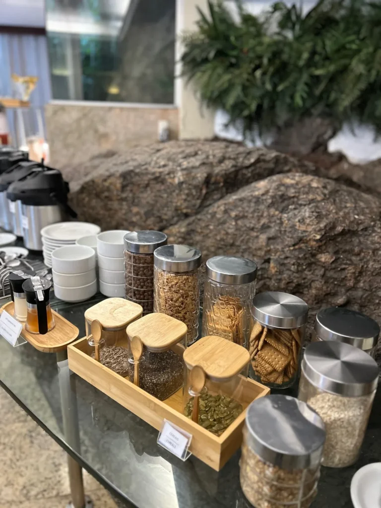 Bancada de cereais, granola, biscoitos e sementes em potes de vidro, com a pedra natural do salão ao fundo