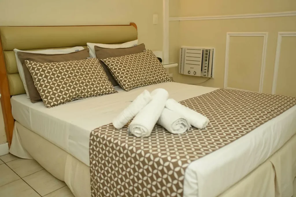 Cama de casal arrumada com toalhas enroladas e almofadas estampadas no Standard Casal