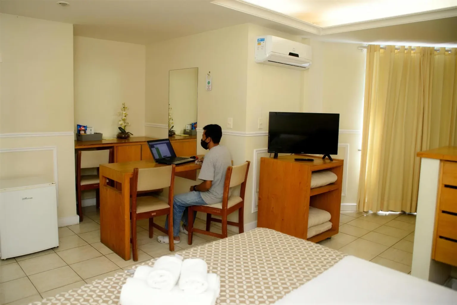 Hóspede trabalhando no notebook na mesa do quarto, com TV e frigobar